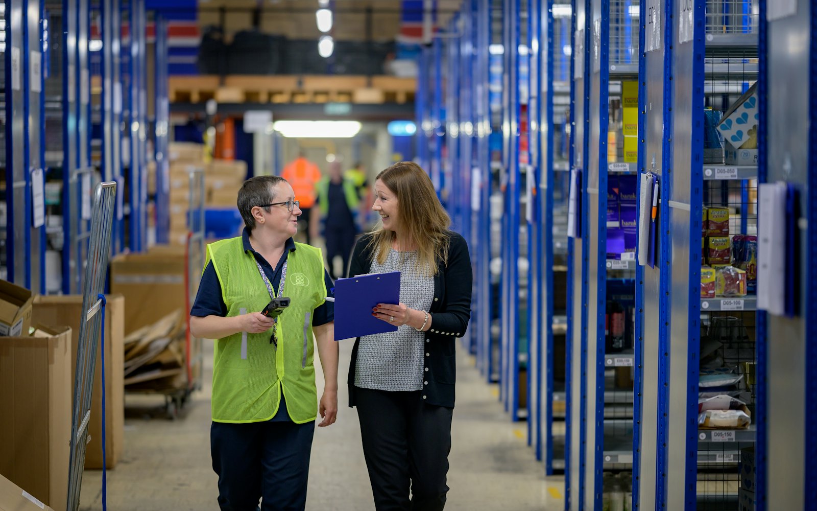 Dos trabajadoras conversan mientras caminan por un pasillo de almacén entre estanterías azules.